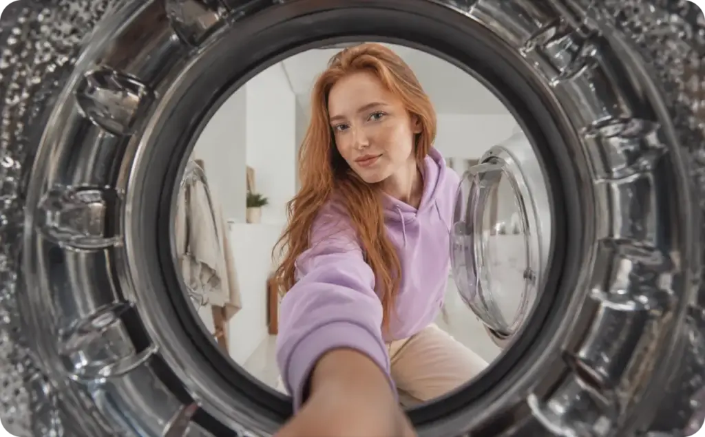 Scroll-stopping hooks: A red-haired woman in a lavender hoodie reaches into a front-loading washing machine, viewed from inside the drum, creating a bold first-person perspective in a bright modern laundry room.