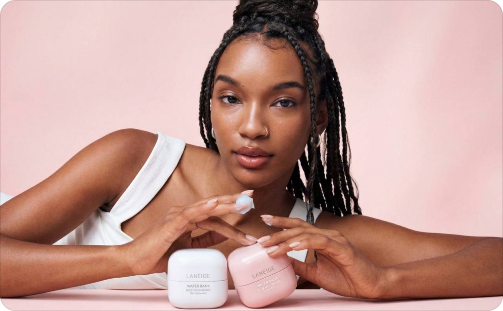 Model on a light pink background looking directly at the camera while touching three skincare creams, illustrating Brand-created ads vs AI UGC ads in a beauty campaign setting