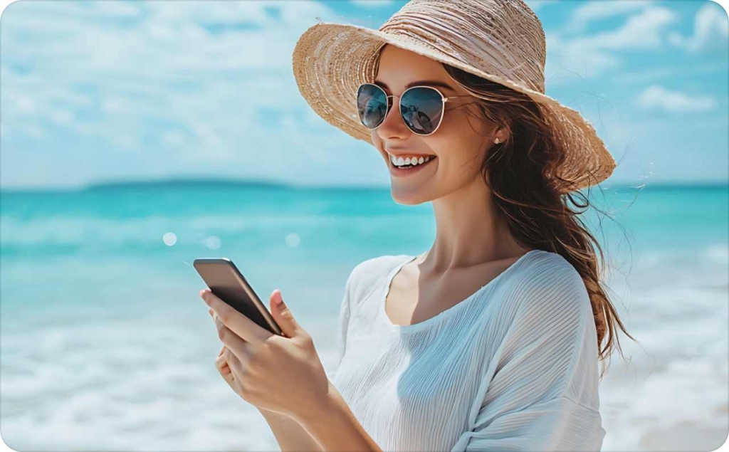 Woman smiling on a sunny beach in a straw hat and sunglasses, holding her phone—perfect for creating beach Instagram captions with summer vibes and sunlight.