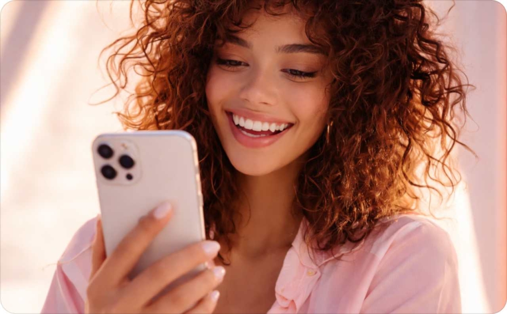 Smiling young woman with curly hair looking at her smartphone, feeling inspired while browsing funny caption ideas on her phone.