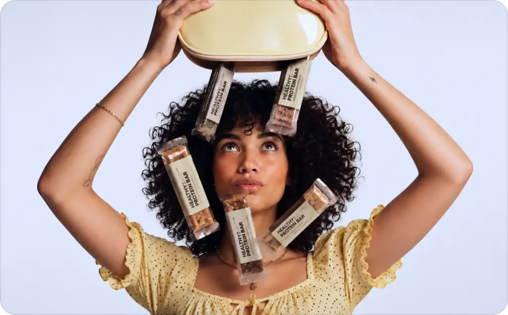 Woman with curly hair in a yellow polka-dot blouse holds a container above her head while several healthy protein bars fall around her against a clean white background. This playful lifestyle image could be used to promote Free AI avatar video generators, health brands, influencer marketing, social media content creation, and digital advertising campaigns.
